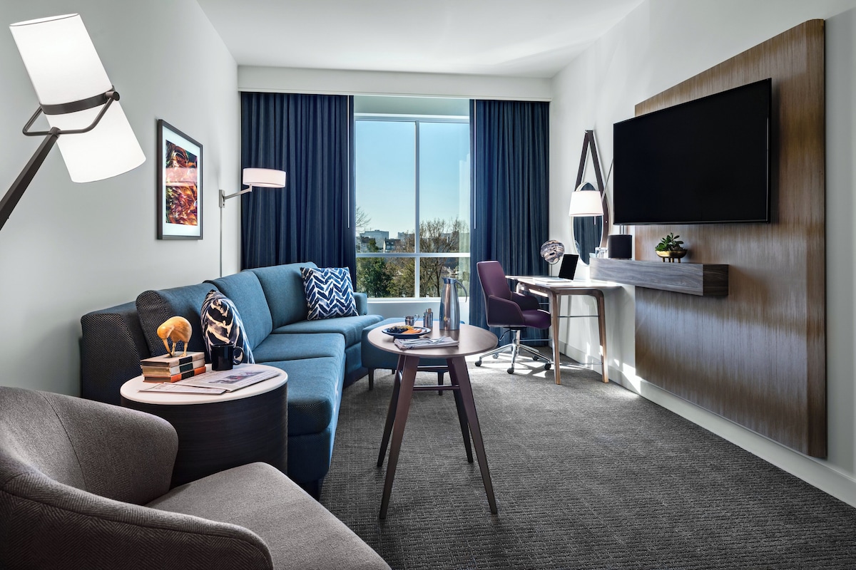A modern hotel room with a sofa, armchair, dining table, and a flat-screen TV mounted on the wall.