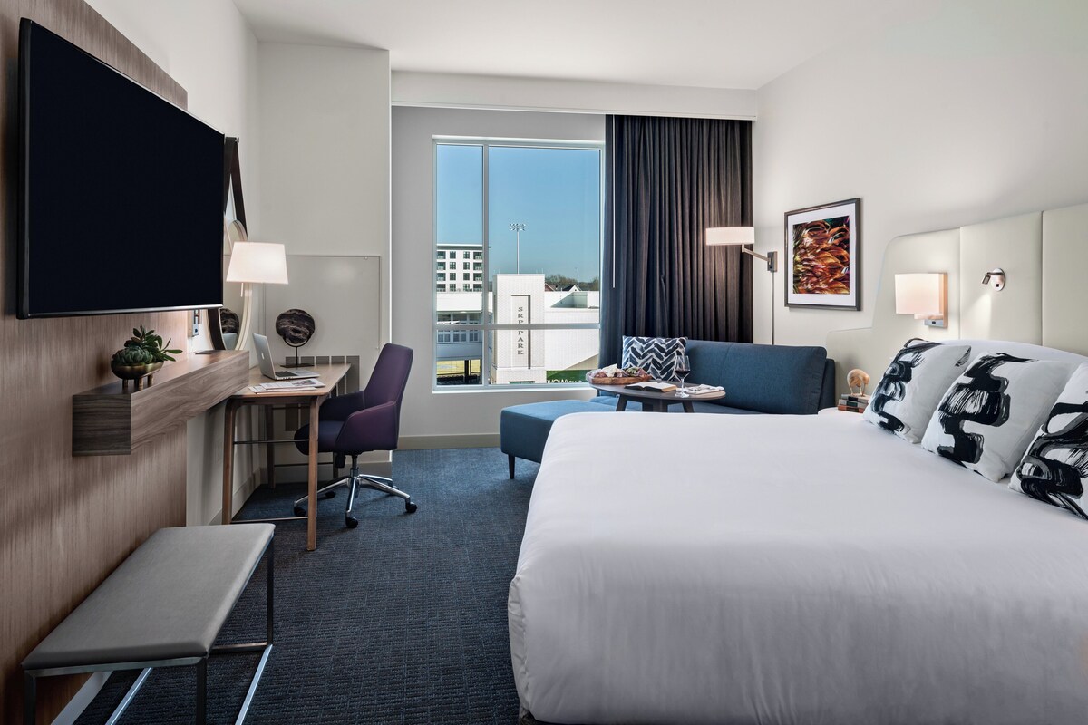 A hotel room with a large bed, a desk with a chair, a small table, a TV mounted on the wall, and a window with a view of buildings.