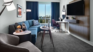 A modern hotel room with a sofa, armchair, round table, and a flat-screen TV mounted on the wall.