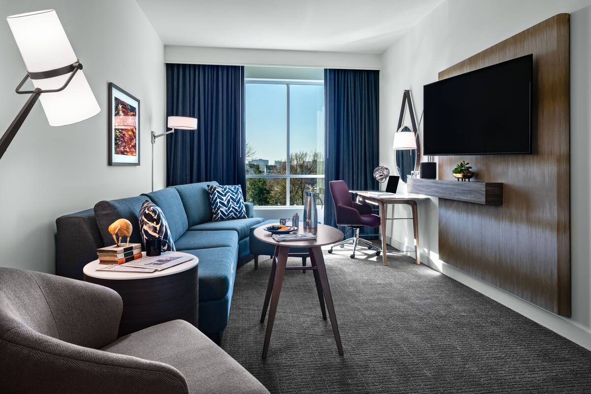Ein modernes Hotelzimmer mit einer Couch, einem Sessel, einem runden Tisch und einem flachbildfernseher an der Wand.