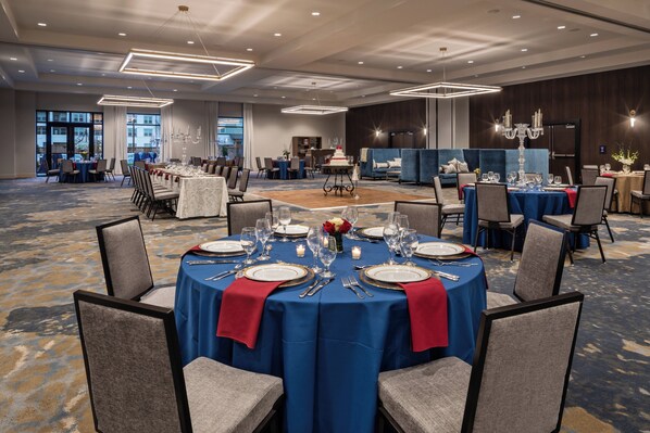 Un salón de banquetes con mesas redondas dispuestas para cenar, una zona de recepción y vistas a la ciudad a través de amplias ventanas.