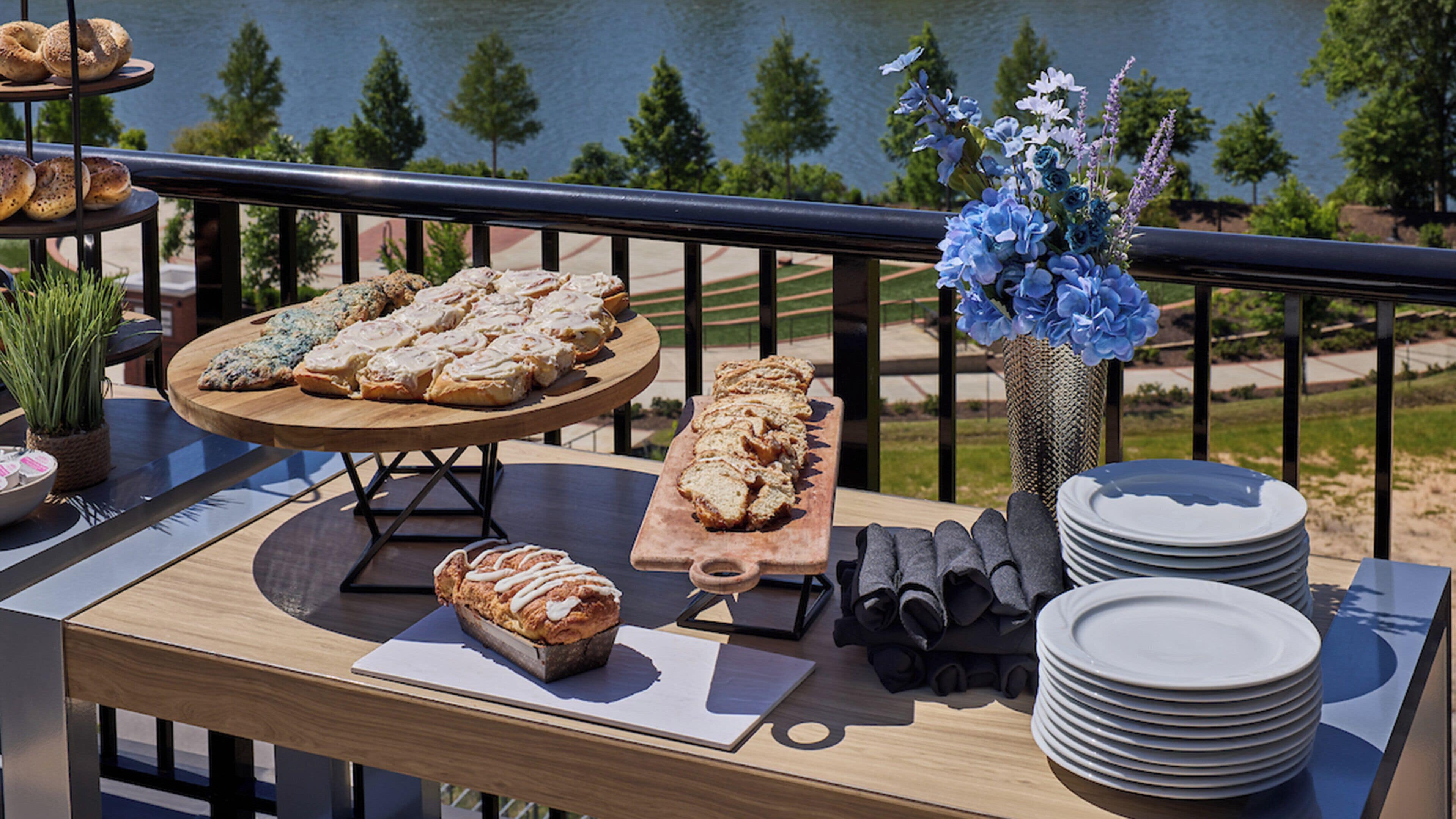 A table with pastries, a vase of flowers, and stacked plates on a balcony with a lake view.
