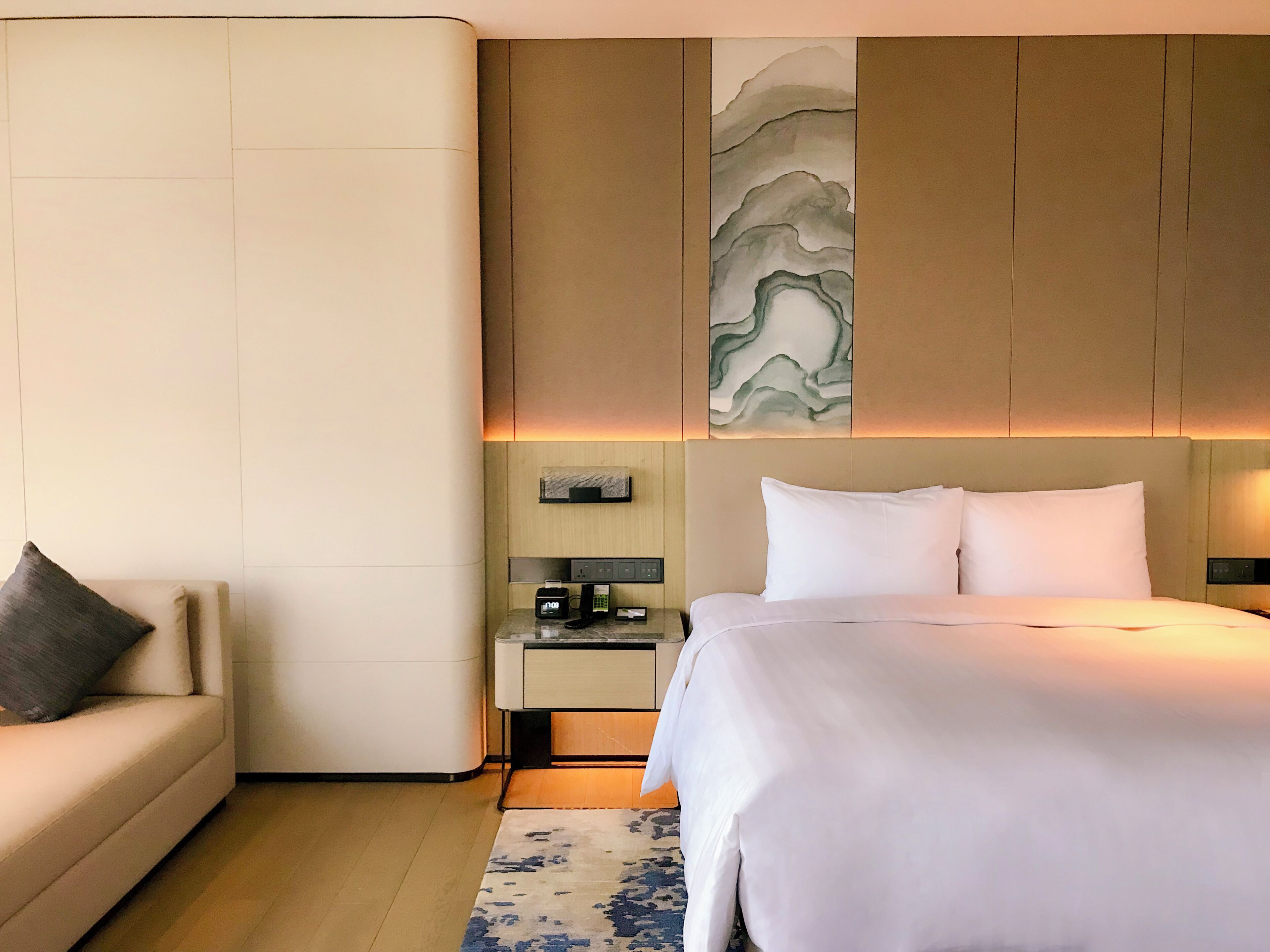 Ein Hotelzimmer mit Bett, Nachttisch, Lampe, Wandbild und einer Couch.