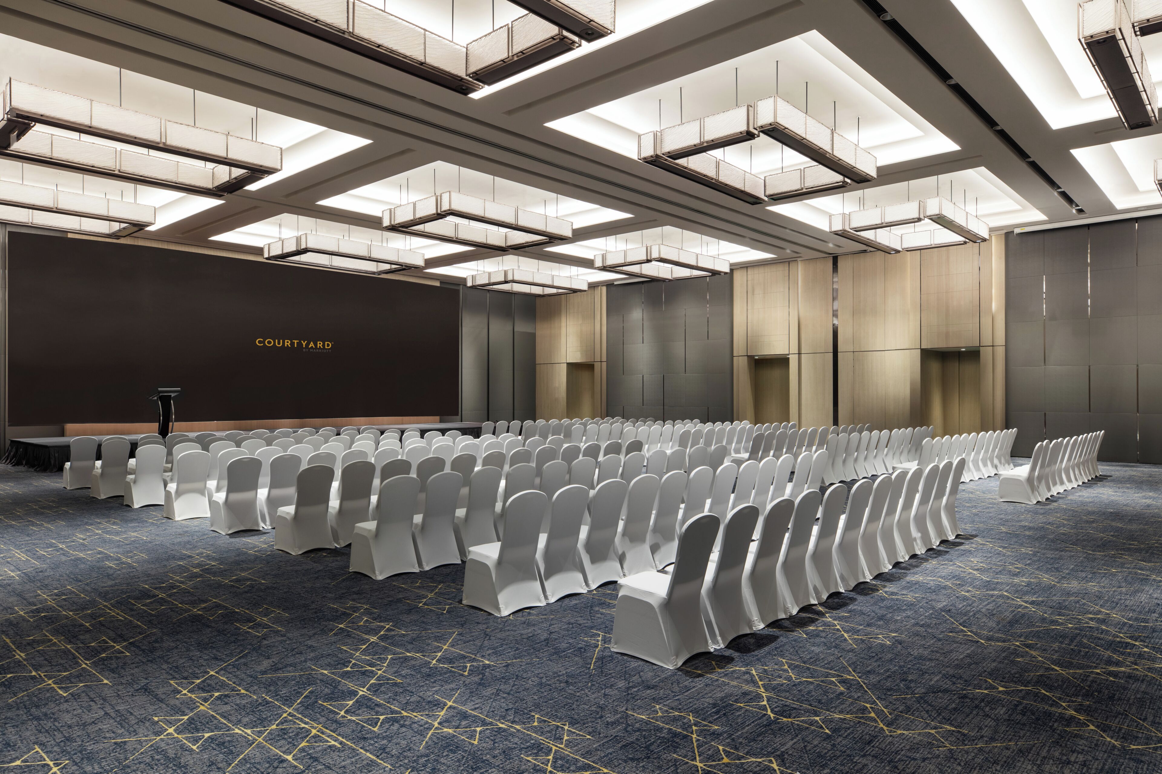 A conference room with rows of chairs, a stage, and a large screen.