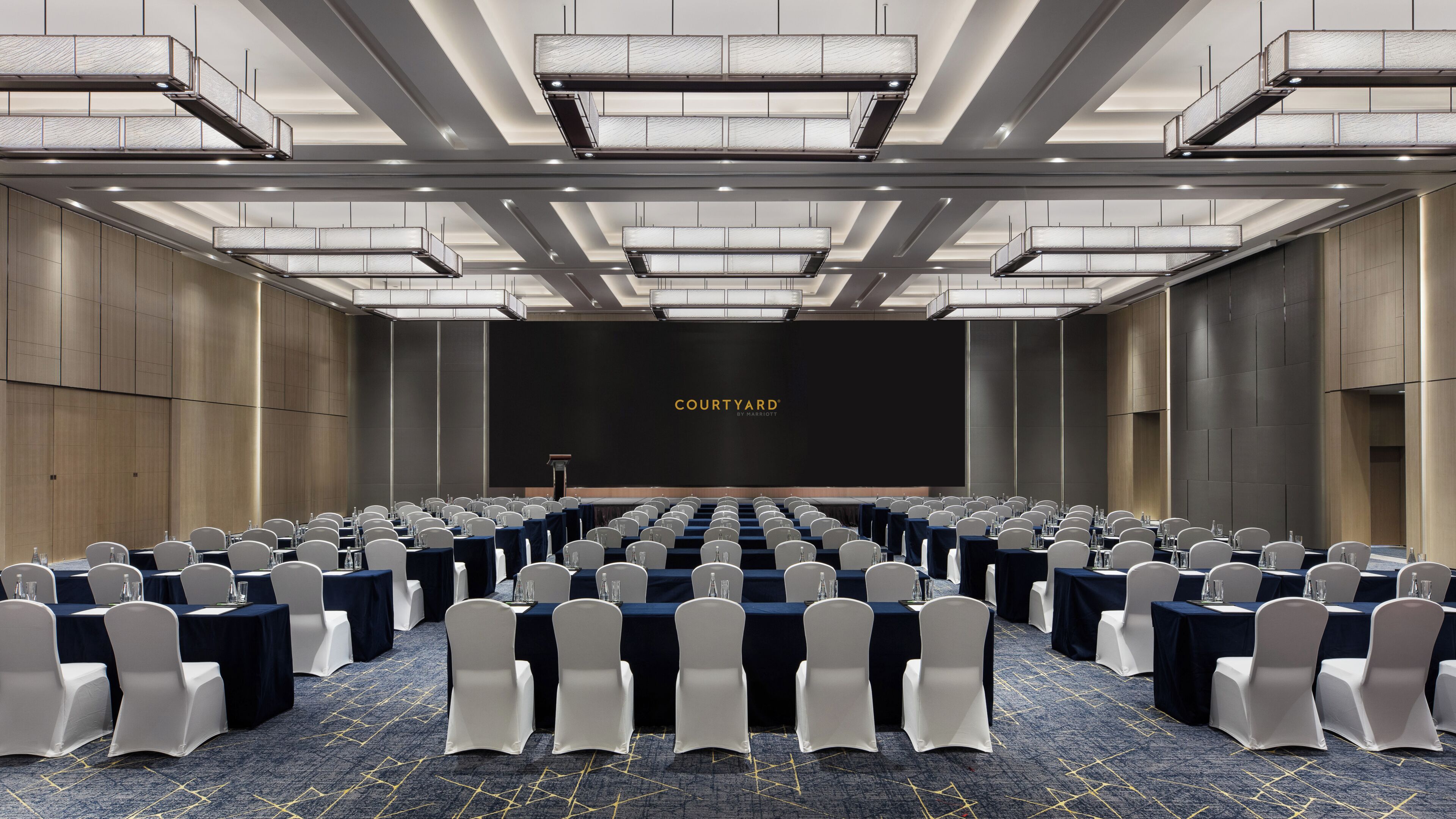 A conference room with rows of tables and chairs, a podium, and a large screen displaying 'COURTYARD BY MARIEOTTA'.
