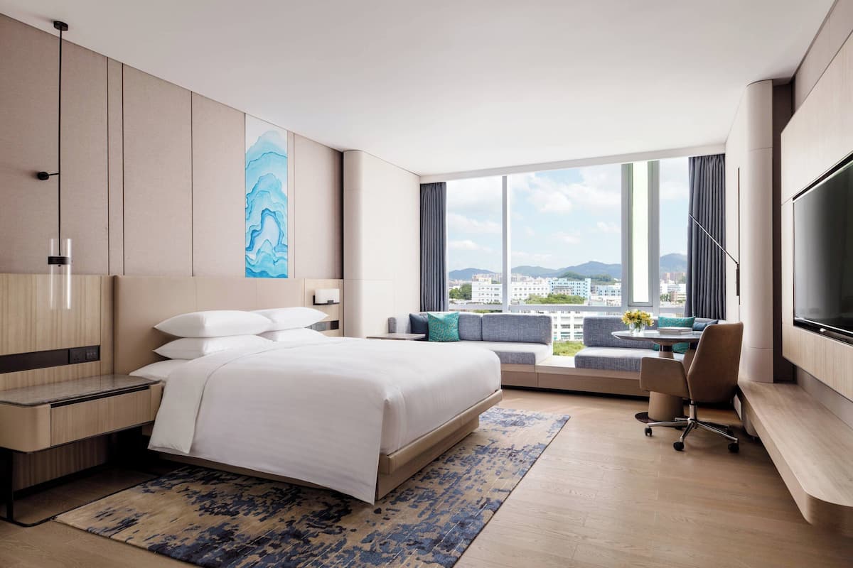 Een moderne hotelkamer met een groot bed, een bureau, een stoel, een televisie en stadszicht door grote ramen.