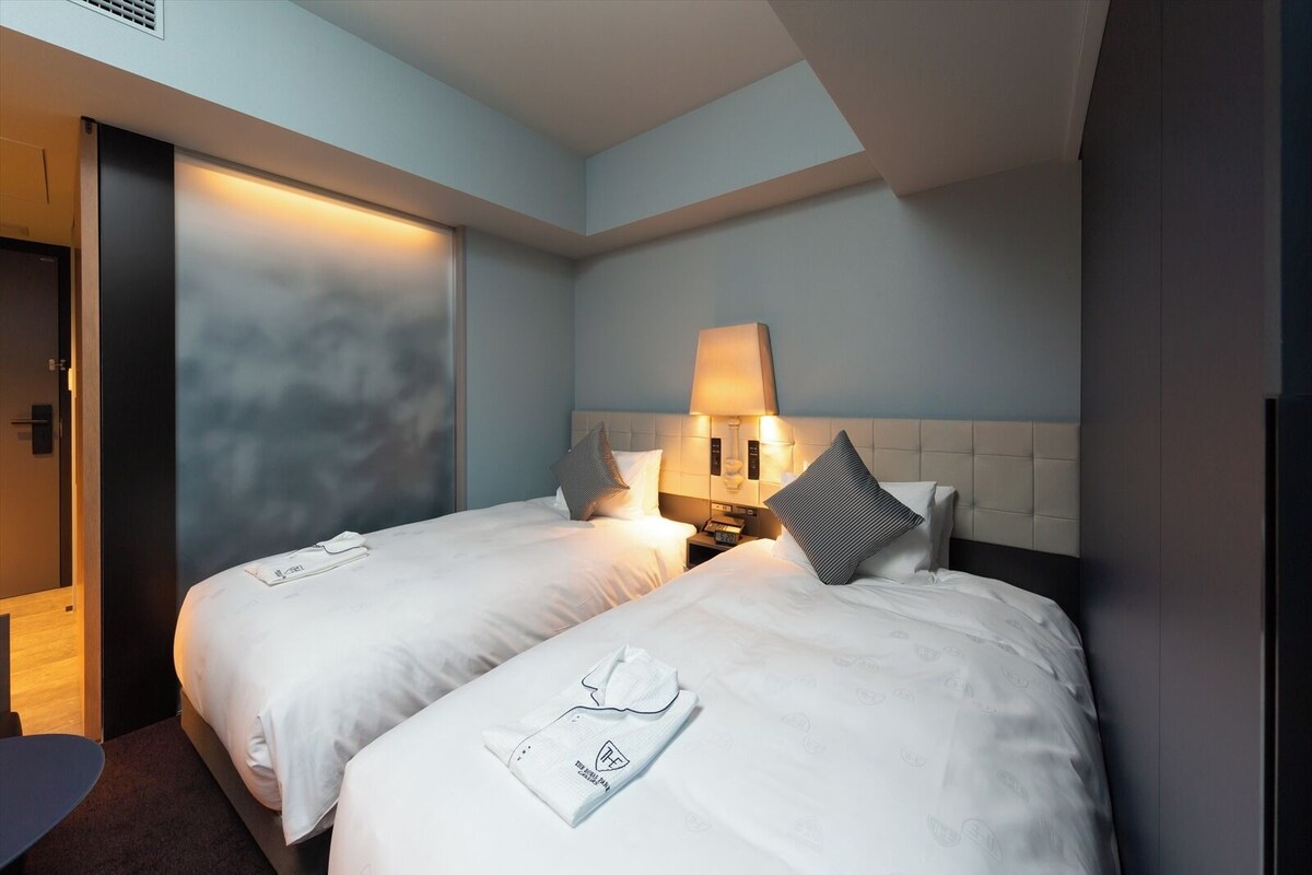 Een hotelkamer met twee bedden, een nachtkastje met een lamp, een tv aan de muur en een deur die naar een andere kamer leidt.