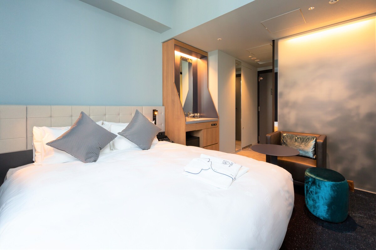 Hotelkamer met een groot bed, nachtkastje, spiegel, zithoek en een klein rond poef.