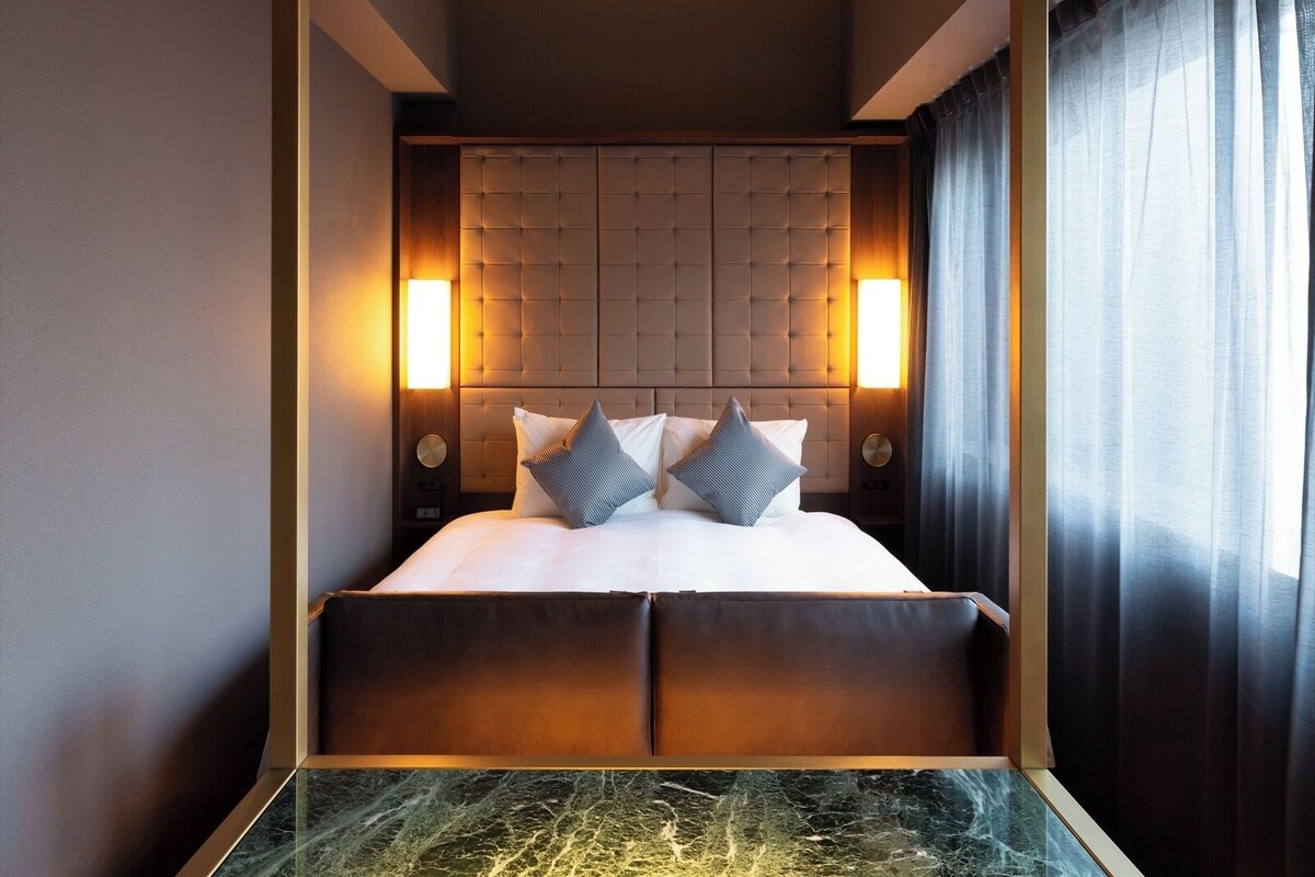 Een hotelkamer met een gestuikt hoofdeinde, twee bedlampen, een bed met witte lakens en een nachtkastje van marmer.