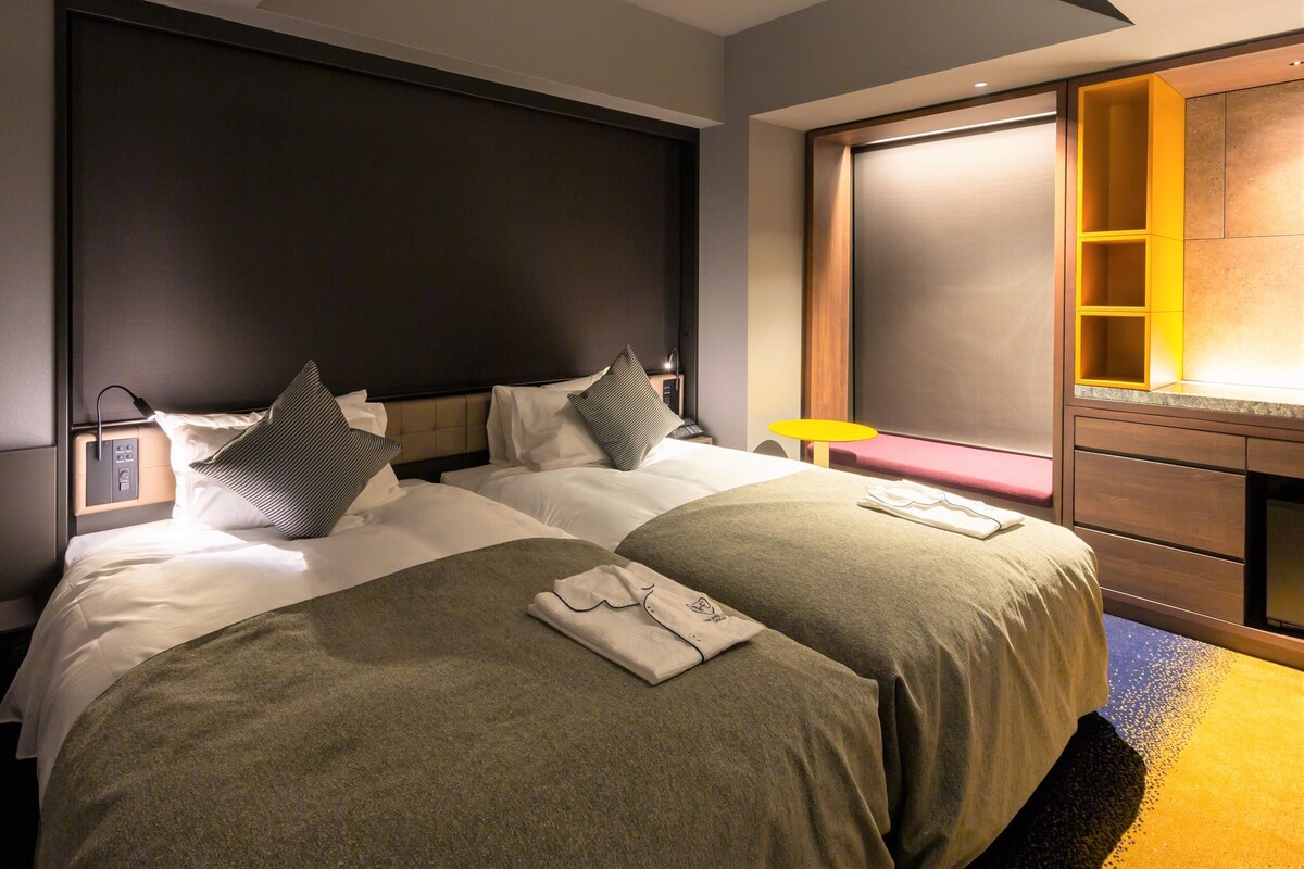 Een hotelkamer met twee bedden, een nachtkastje, een bureau met stoel, een groot raam en een inbouwkast.