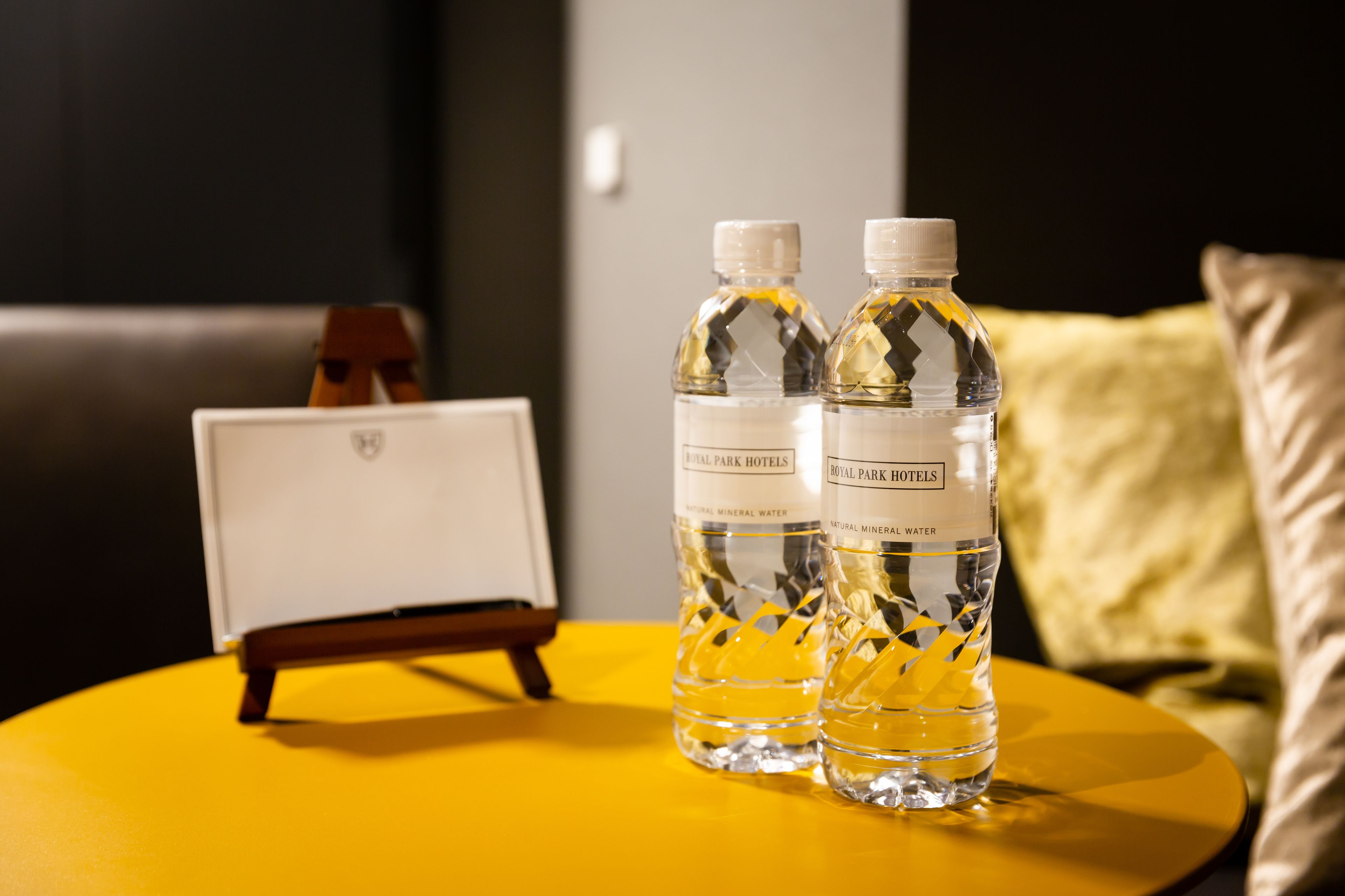 Two bottles of Royal Park Hotels mineral water on a yellow table with a framed picture and a yellow pillow in the background.