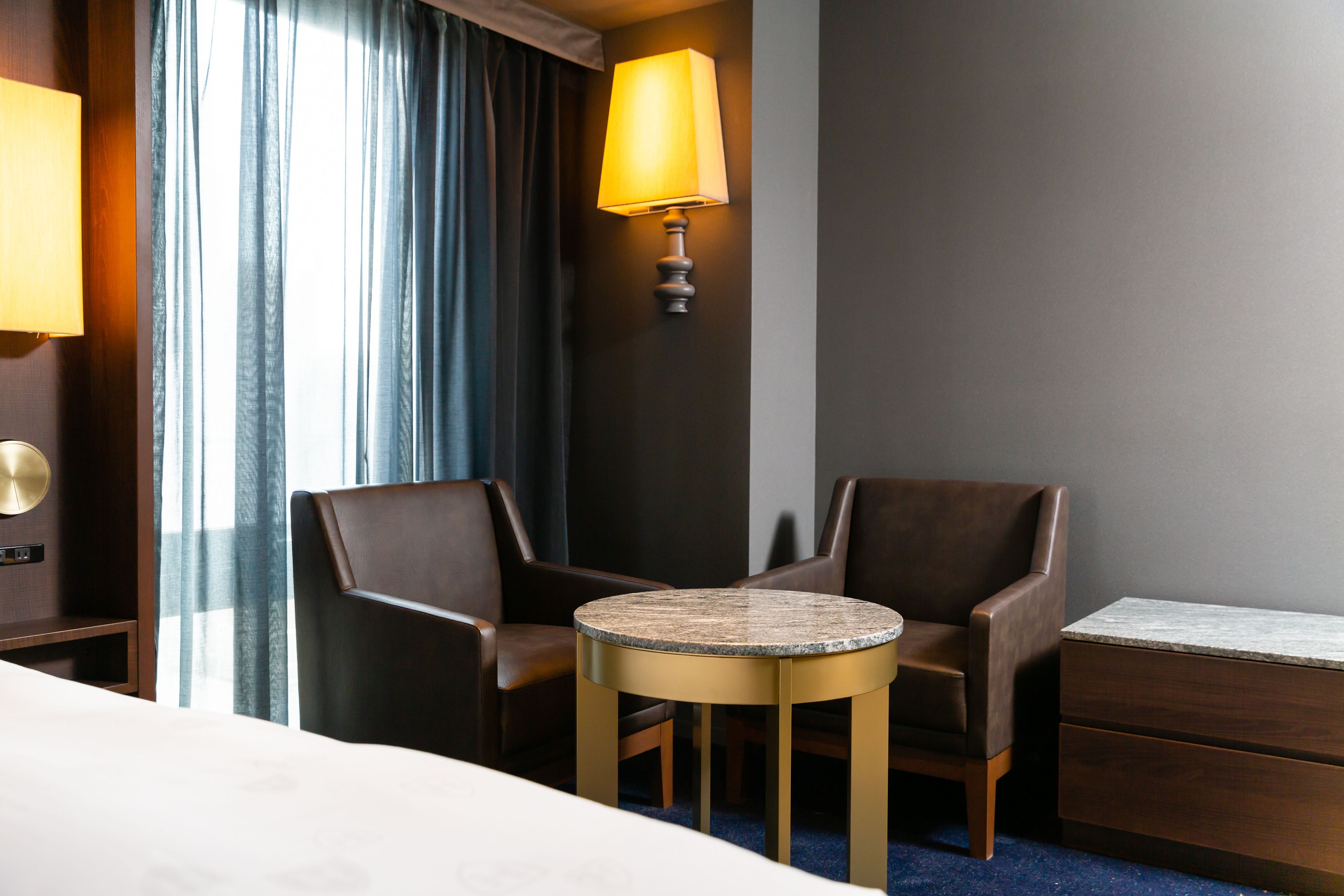 A hotel room with a bed, two armchairs, a small round table, a lamp, and a nightstand.