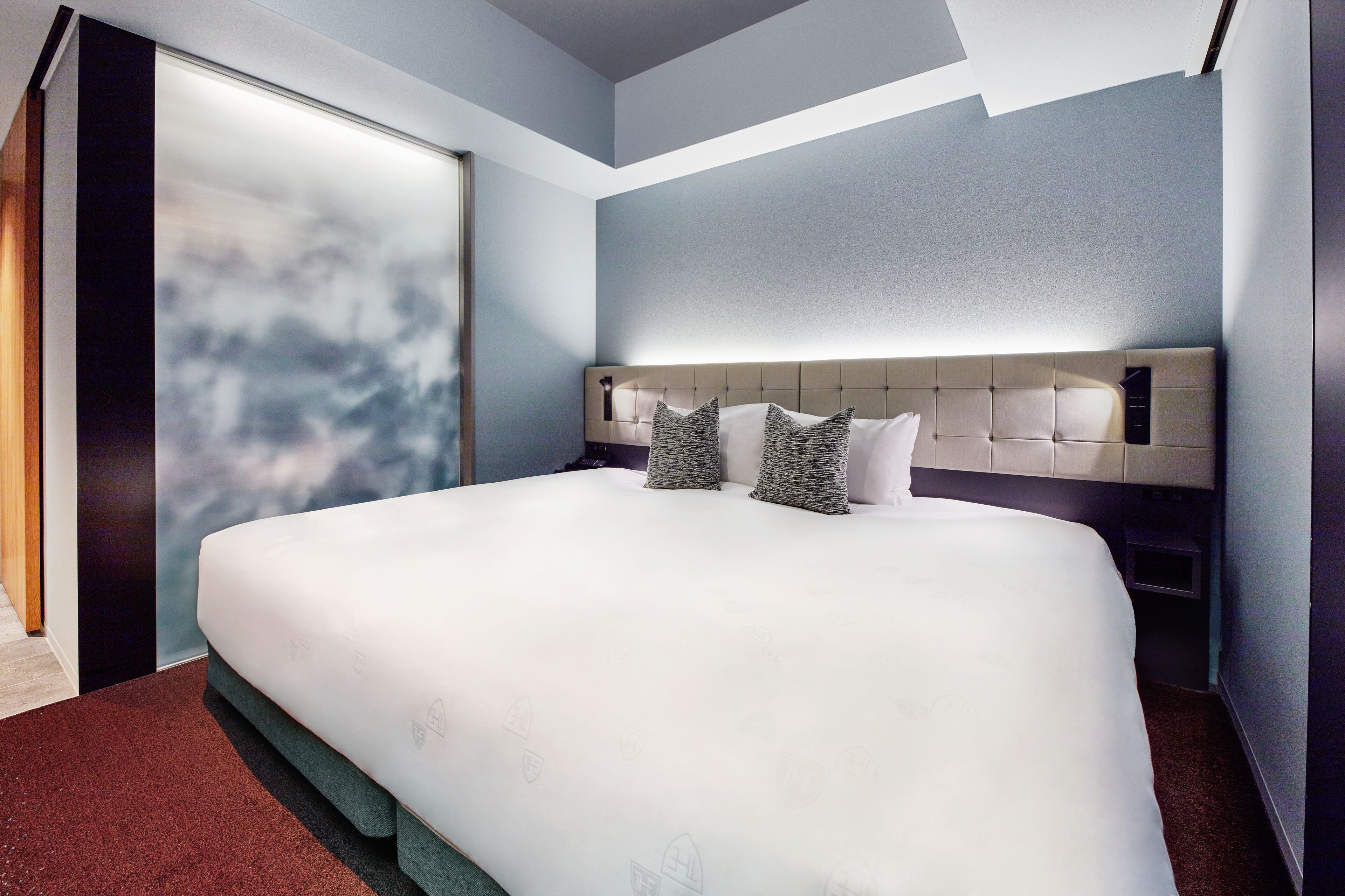 A modern hotel room with a large bed, a headboard with built-in controls, and a large window with a view of the sky.