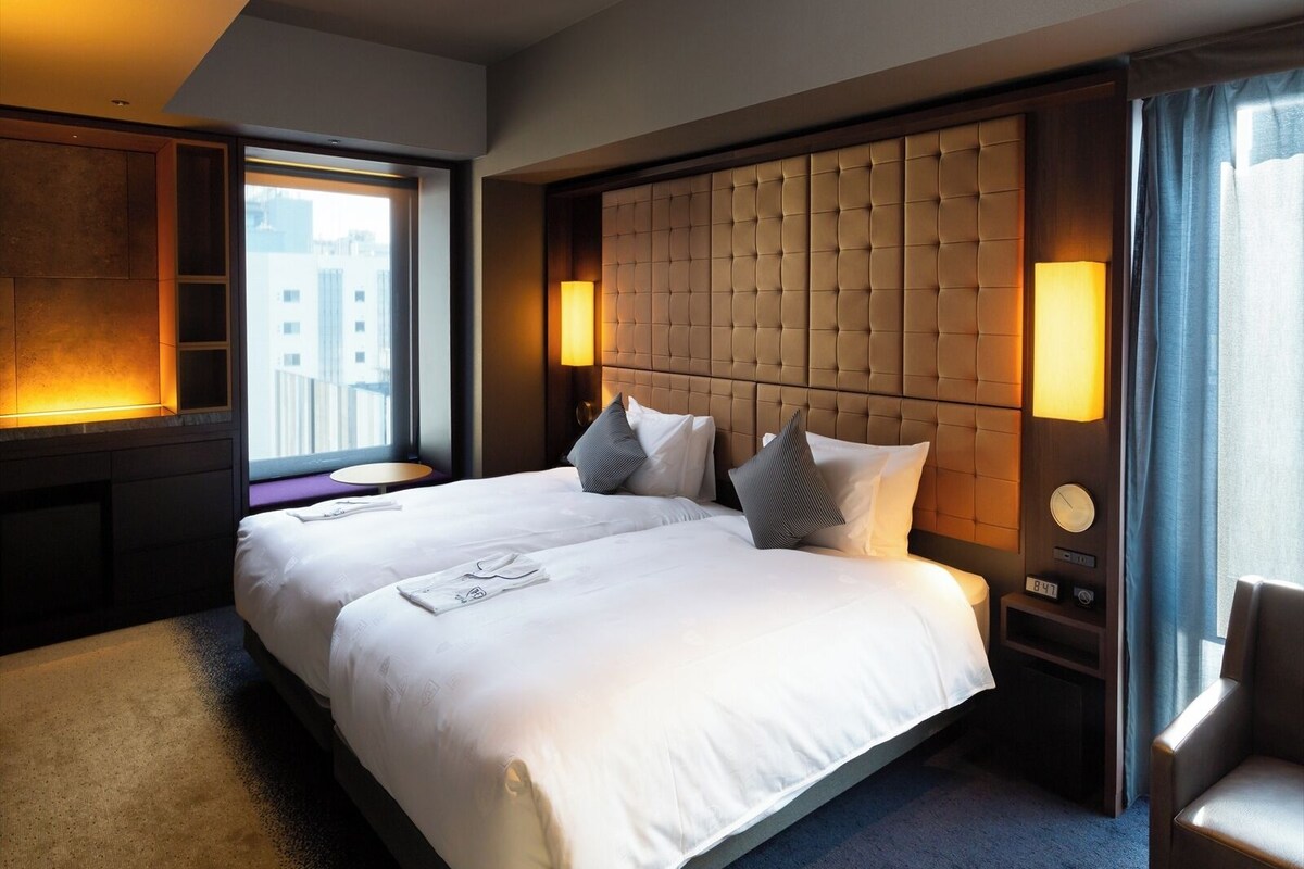Hotelkamer met een groot bed, nachtkastje, stoel en raam met uitzicht over de stad.