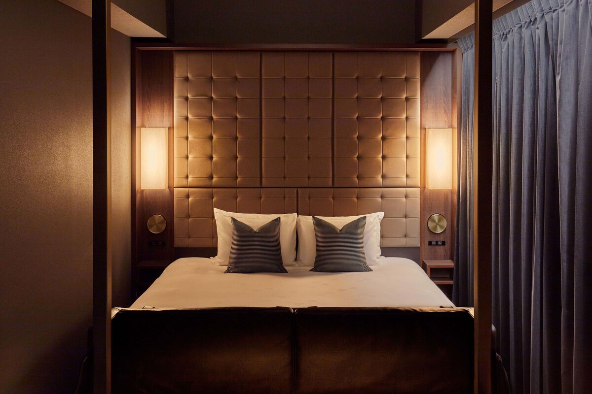Een hotelkamer met een gestoffeerd hoofdeinde, een bed met twee kussens en een nachtkastje met een lamp.