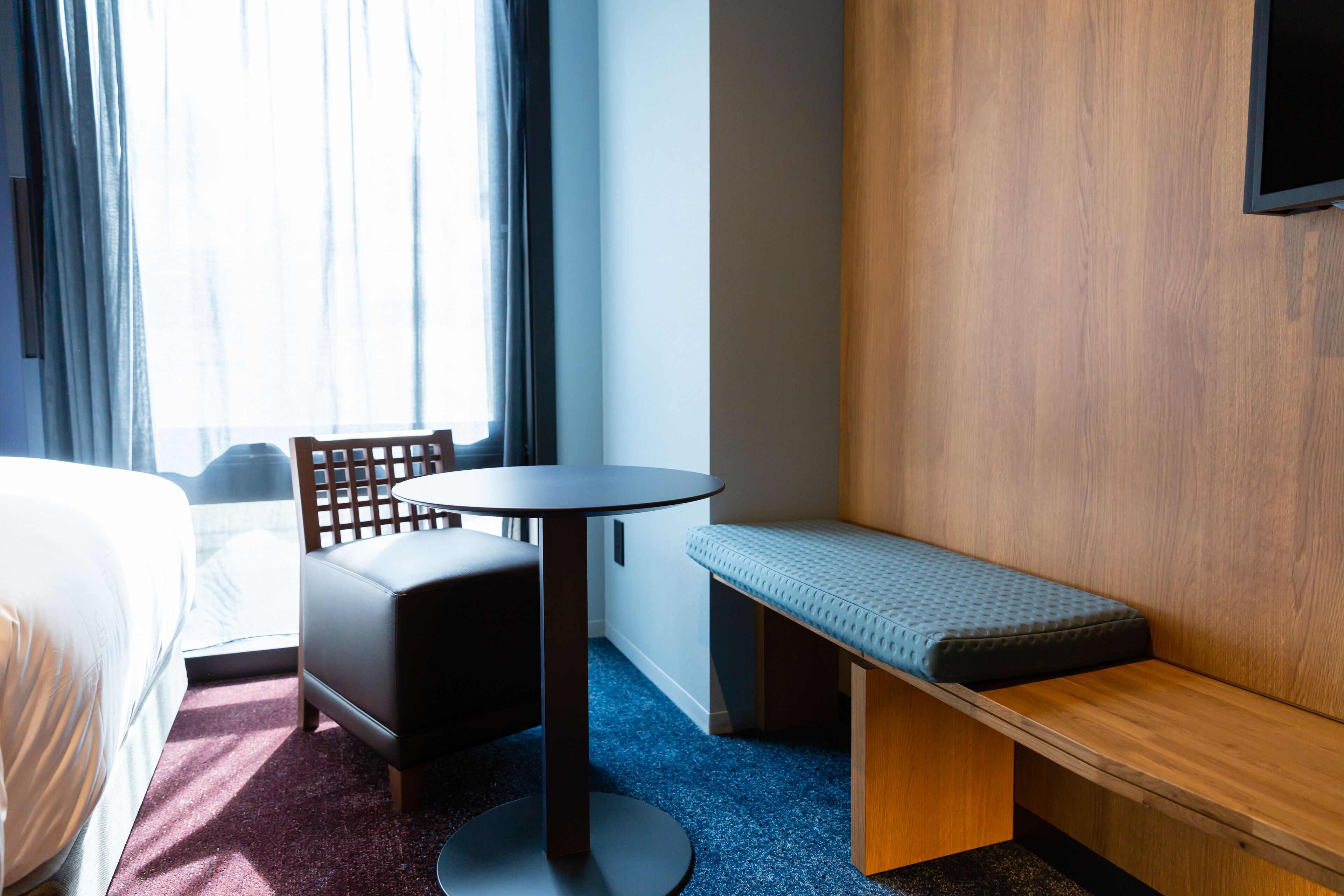 A hotel room with a bed, a round table, a bench, and a TV mounted on the wall.
