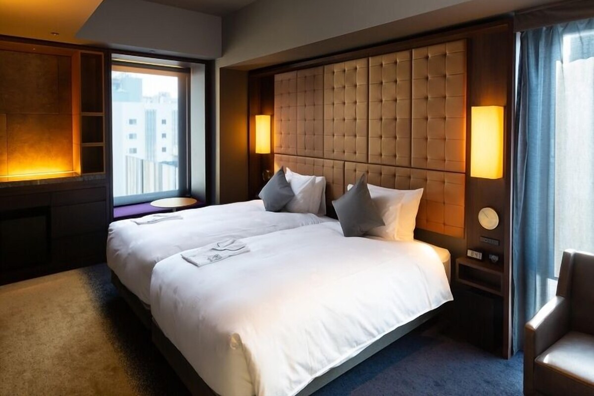Hotelkamer met een groot bed, hoofdbord, bedlampen, een fauteuil en uitzicht op de stad.