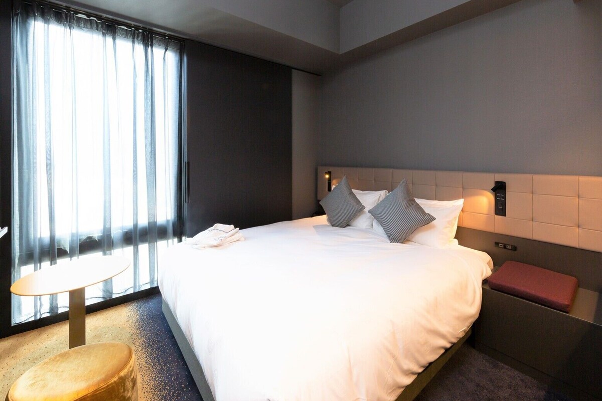 Een hotelkamer met een groot bed, nachtkastje, lamp en een klein krukje.