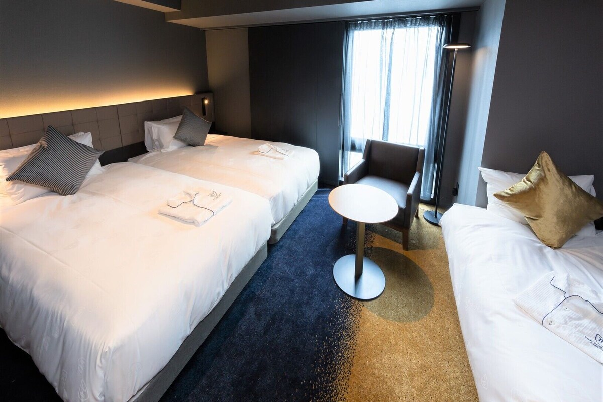 Een hotelkamer met twee bedden, een klein rond tafeltje, een stoel en een groot raam met gordijnen.