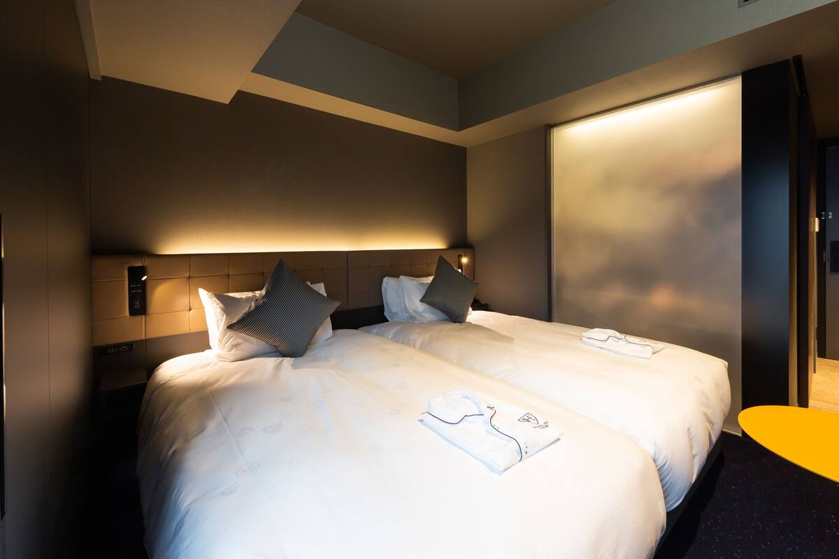 Hotellihuone, jossa on kaksi sänkyä, yöpöytä ja pieni keltainen pöytä.