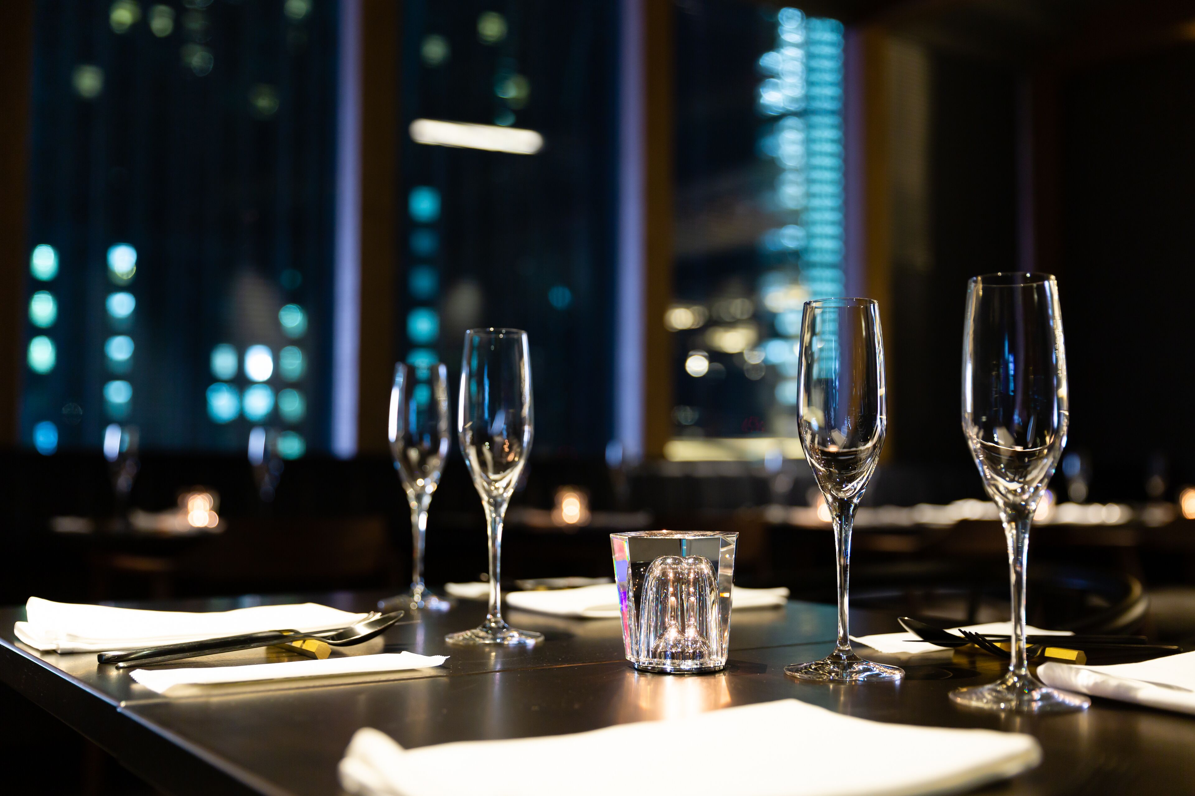 A dining table with four wine glasses and a small glass, set with white napkins and cutlery, against a backdrop of a cityscape at night.