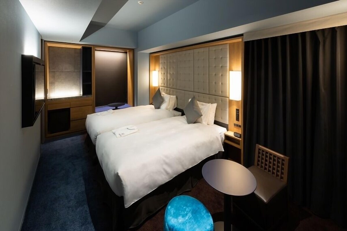 Een hotelkamer met twee bedden, een kleine tafel, een blauwe kruk, een televisie en een groot raam met gordijnen.