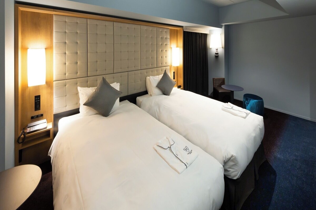 Een hotelkamer met twee bedden, een nachtkastje met een lamp, een klein rond tafeltje en een blauwe stoel.