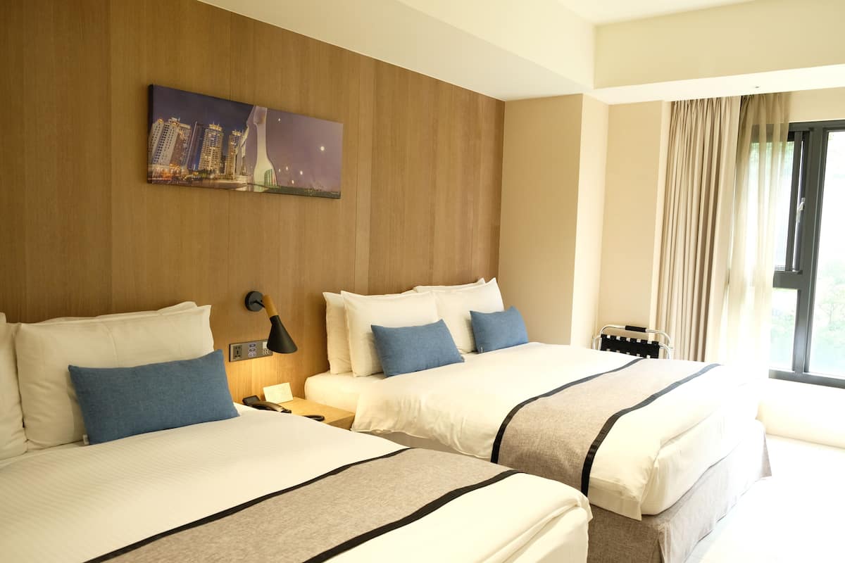 A hotel room with two beds, a nightstand, a lamp, a painting, and a window with curtains.