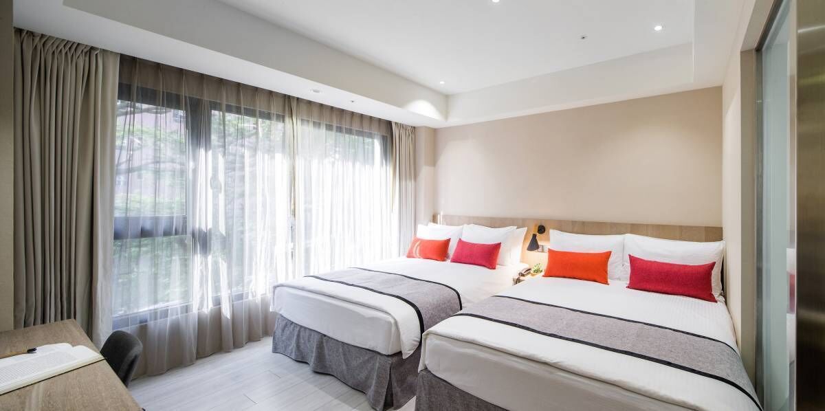 A hotel room with two beds, a desk, and a large window with curtains.