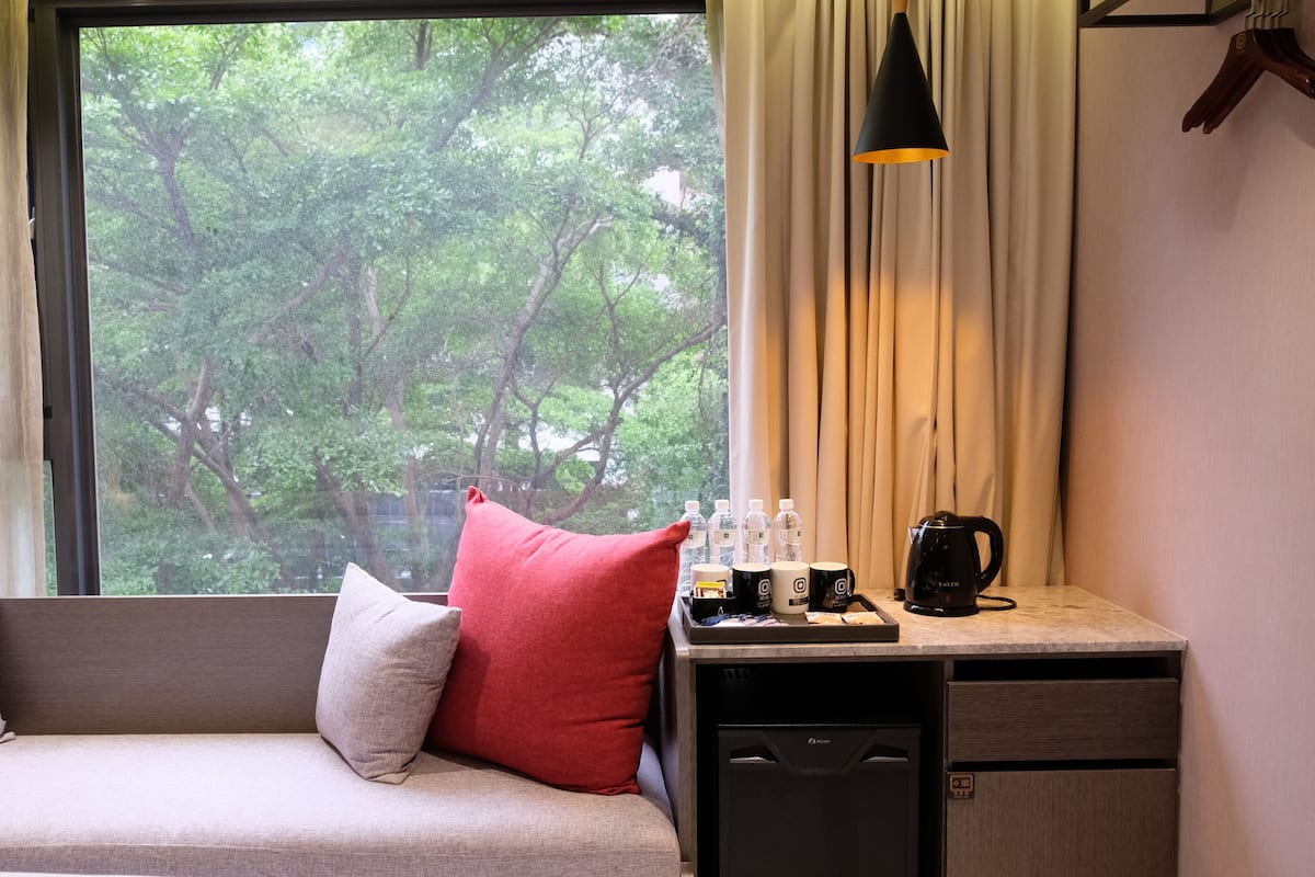 A room with a sofa, pillows, a small table with a coffee maker, bottles, and a view of trees through a window.