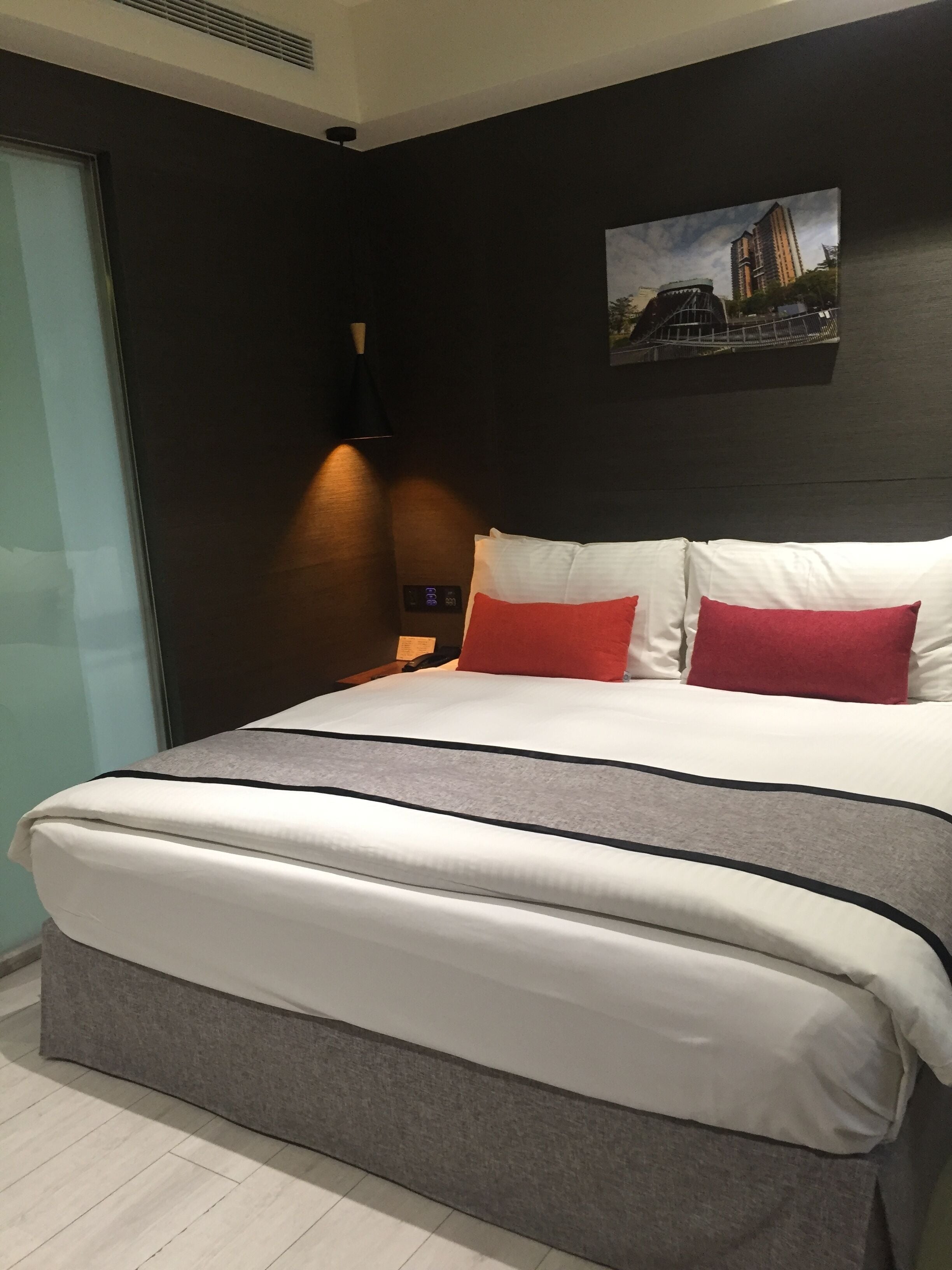 A modern hotel room with a large bed, a digital clock, a painting of a building, and a sliding glass door.