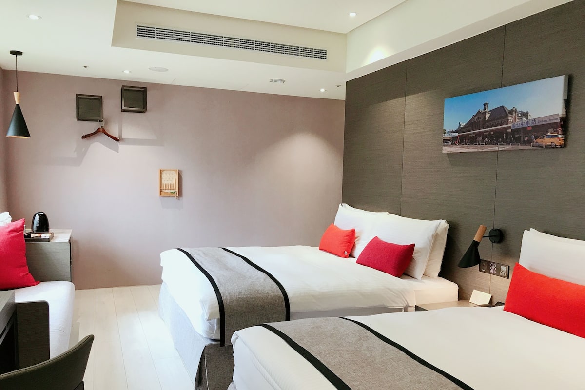 A hotel room with two beds, a desk, a chair, a coffee maker, a hanging light, a wall-mounted clock, and a framed picture.
