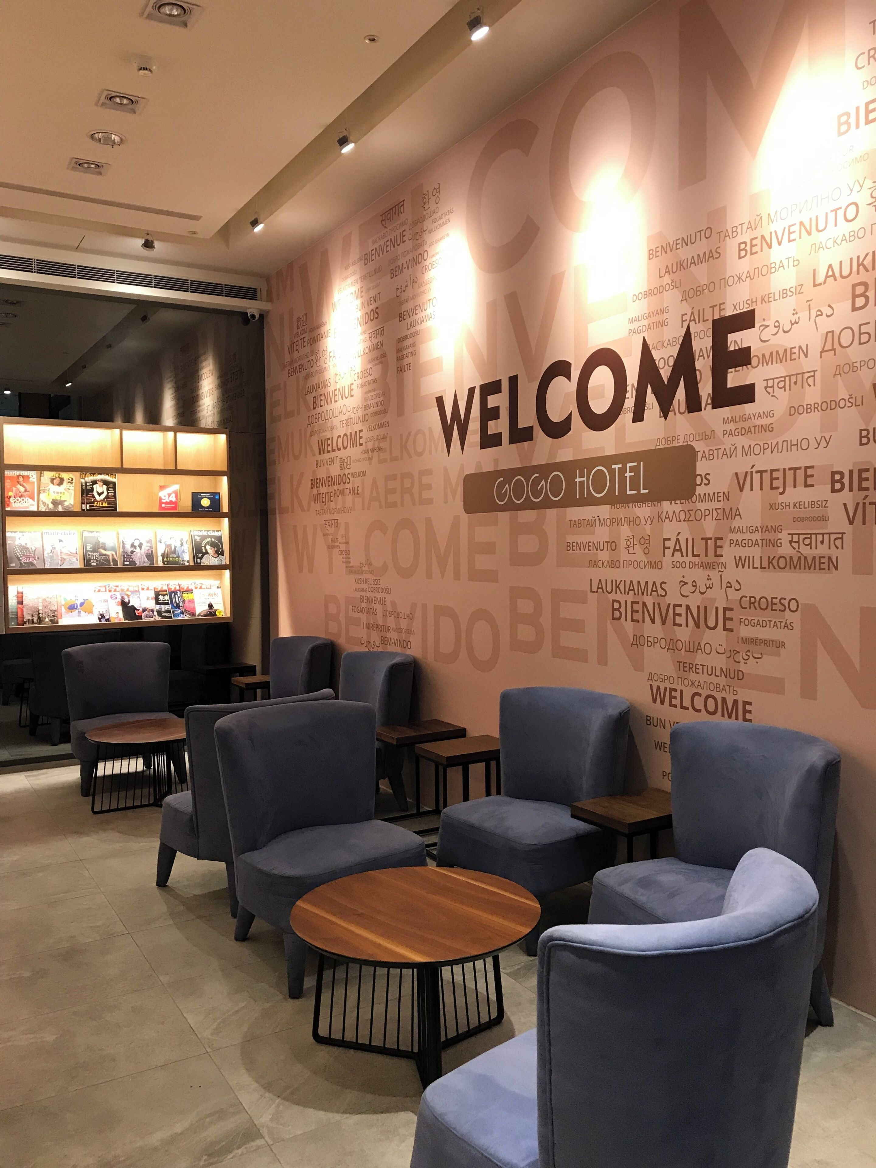 A hotel lobby with multiple seating areas, a reception desk, and a wall displaying the word 'WELCOME' in multiple languages.