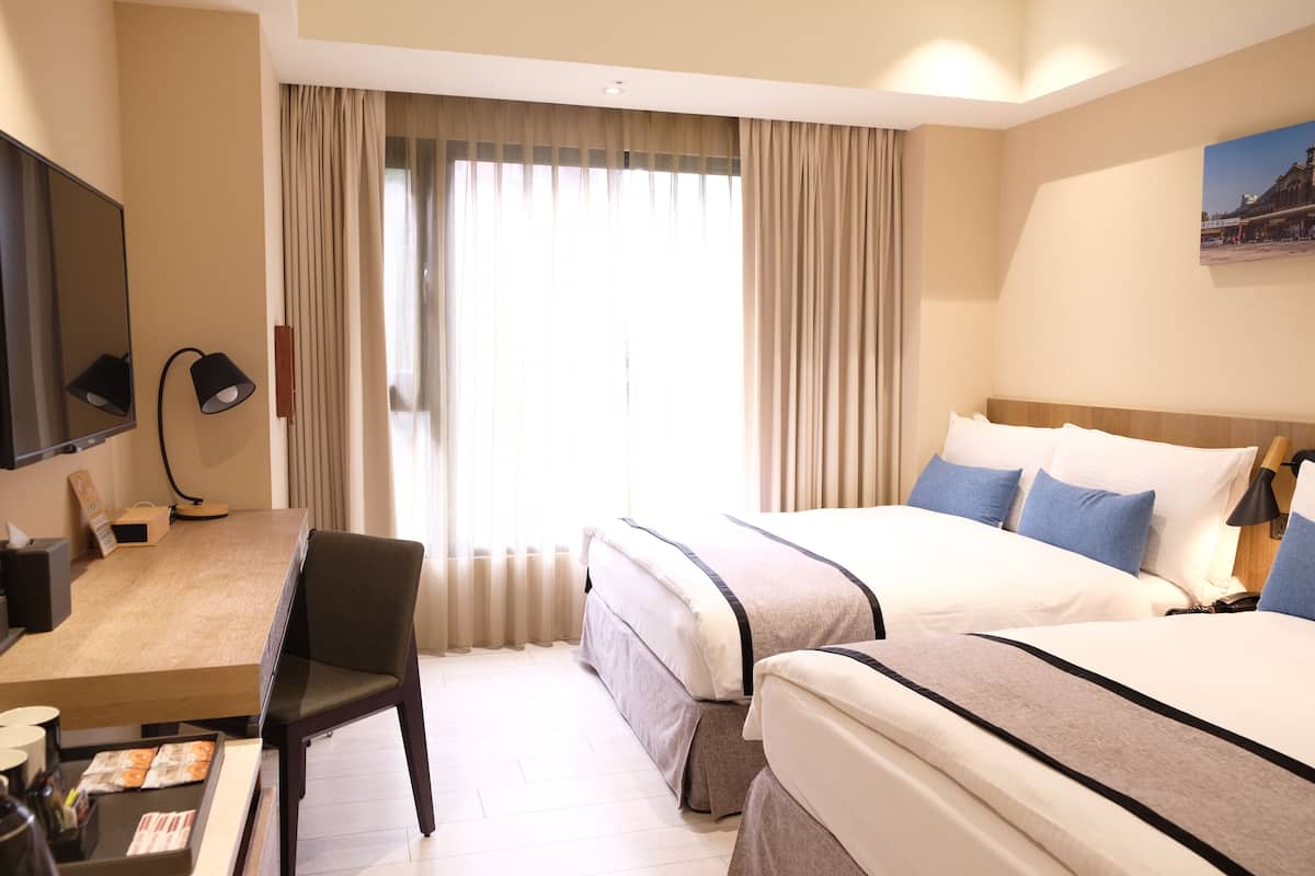 A hotel room with two beds, a desk, a chair, a TV, and a large window with curtains.