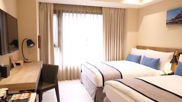 A hotel room with two beds, a desk, a chair, a TV, and a large window with curtains.