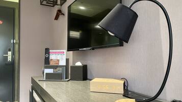 A modern hotel room with a flat-screen TV mounted on the wall, a desk with a clock, a lamp, and a small wooden box.