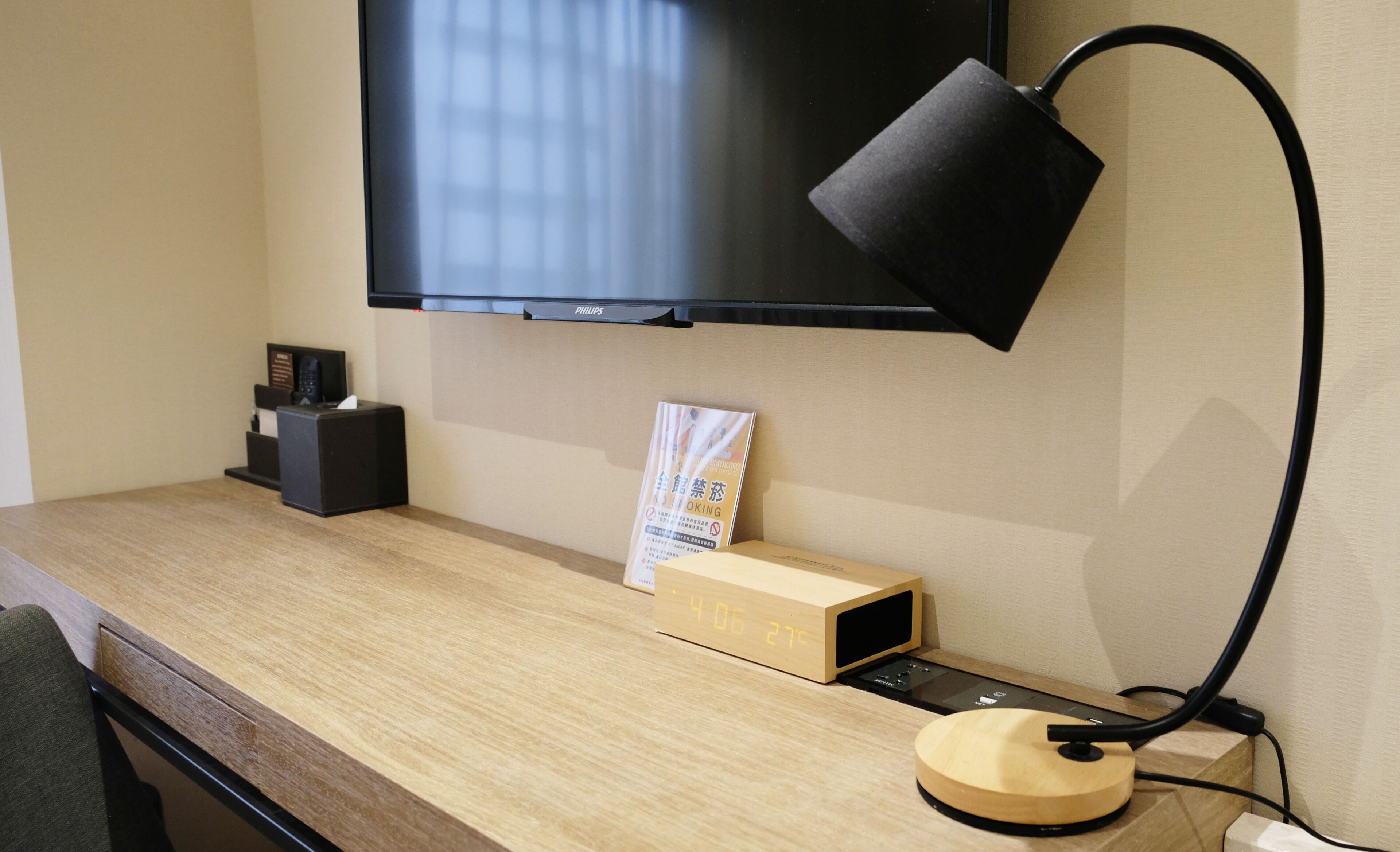 Ein Hotelzimmer mit einem Holzbüro, einem flachbildschirmfernseher an der Wand, einer Schreibtischlampe, einer Taschentuchbox und einer kleinen Holzkiste.