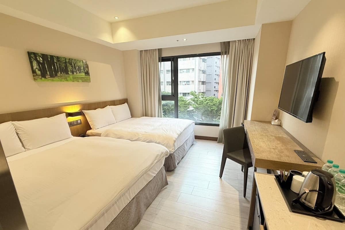 A hotel room with two beds, a desk, a TV, and a window with a view of buildings.