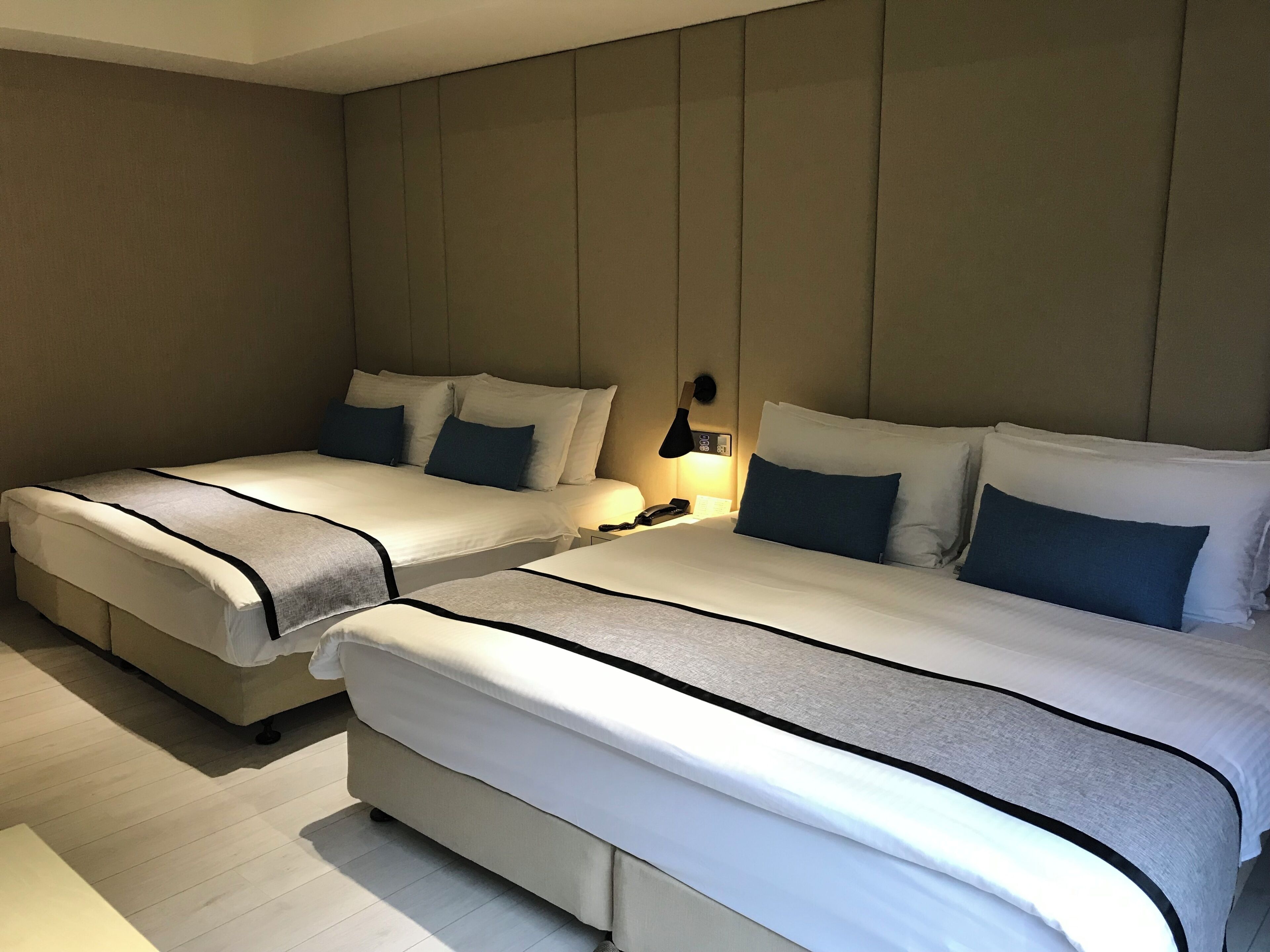 A hotel room with two beds, a bedside table with a lamp, and a wall-mounted phone.