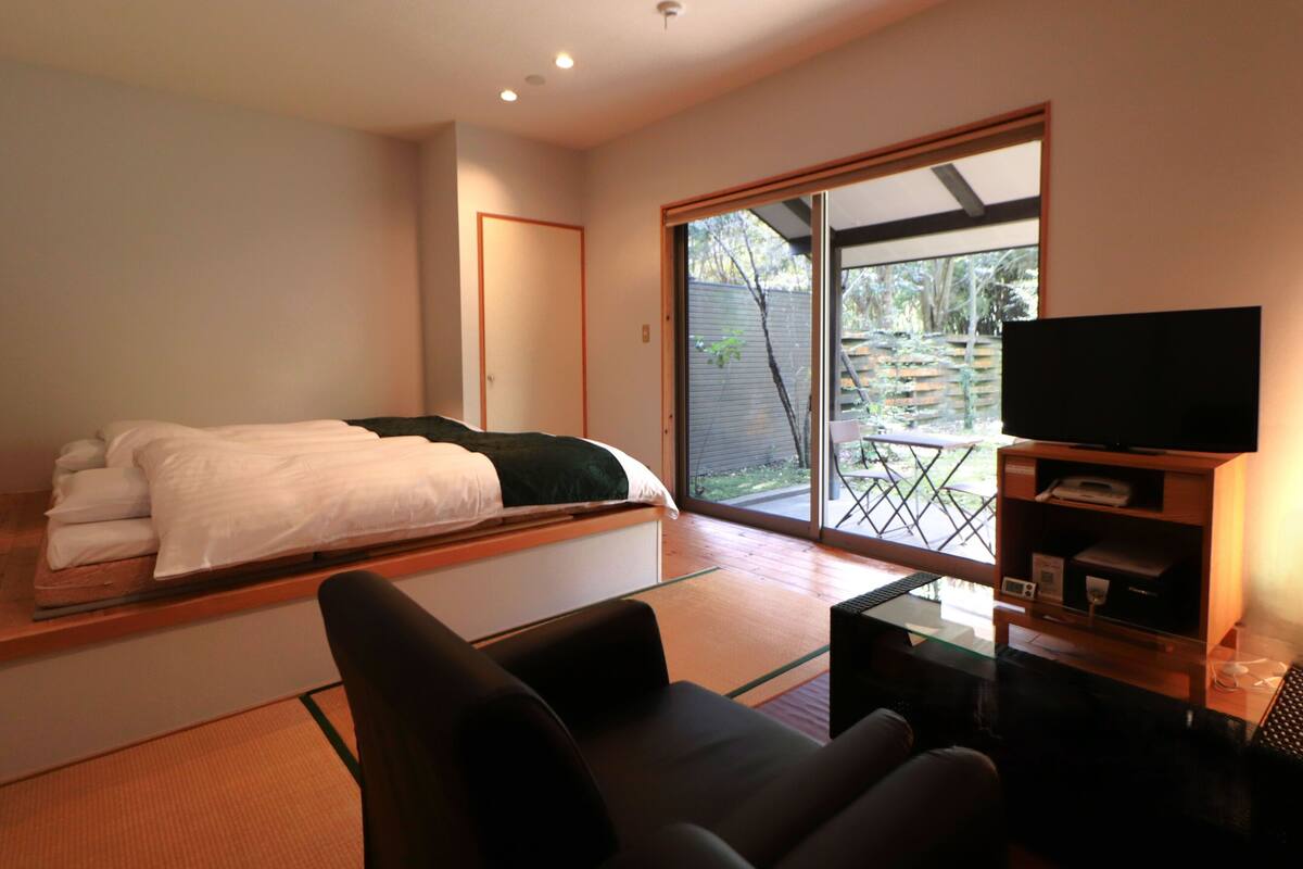 Quarto moderno com cama, sofá, televisão e porta de vidro deslizante para o exterior.