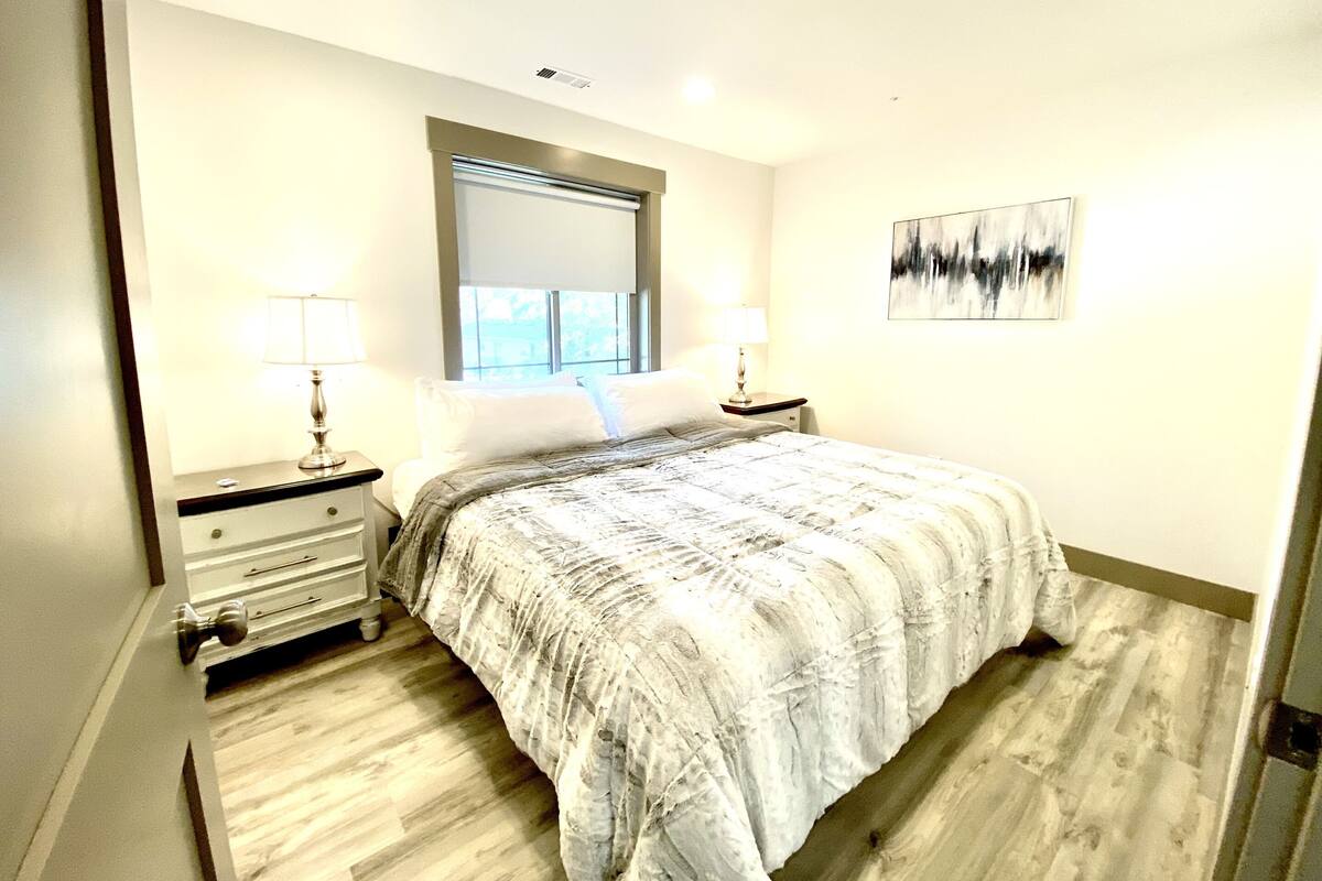 A bedroom with a bed, two bedside tables with lamps, a window with blinds, a wall-mounted artwork, and a wooden floor.