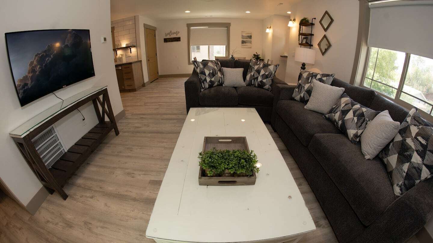 Una sala de estar moderna con un sofá modular, un televisor de pantalla plana montado en la pared, una mesa de centro con una planta y una cocina en el fondo.