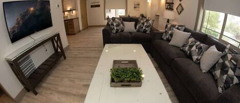 Un salon moderne avec un canapé d’angle, un téléviseur à écran plat fixé au mur, une table basse agrémentée d’une plante, et un coin cuisine en arrière-plan.