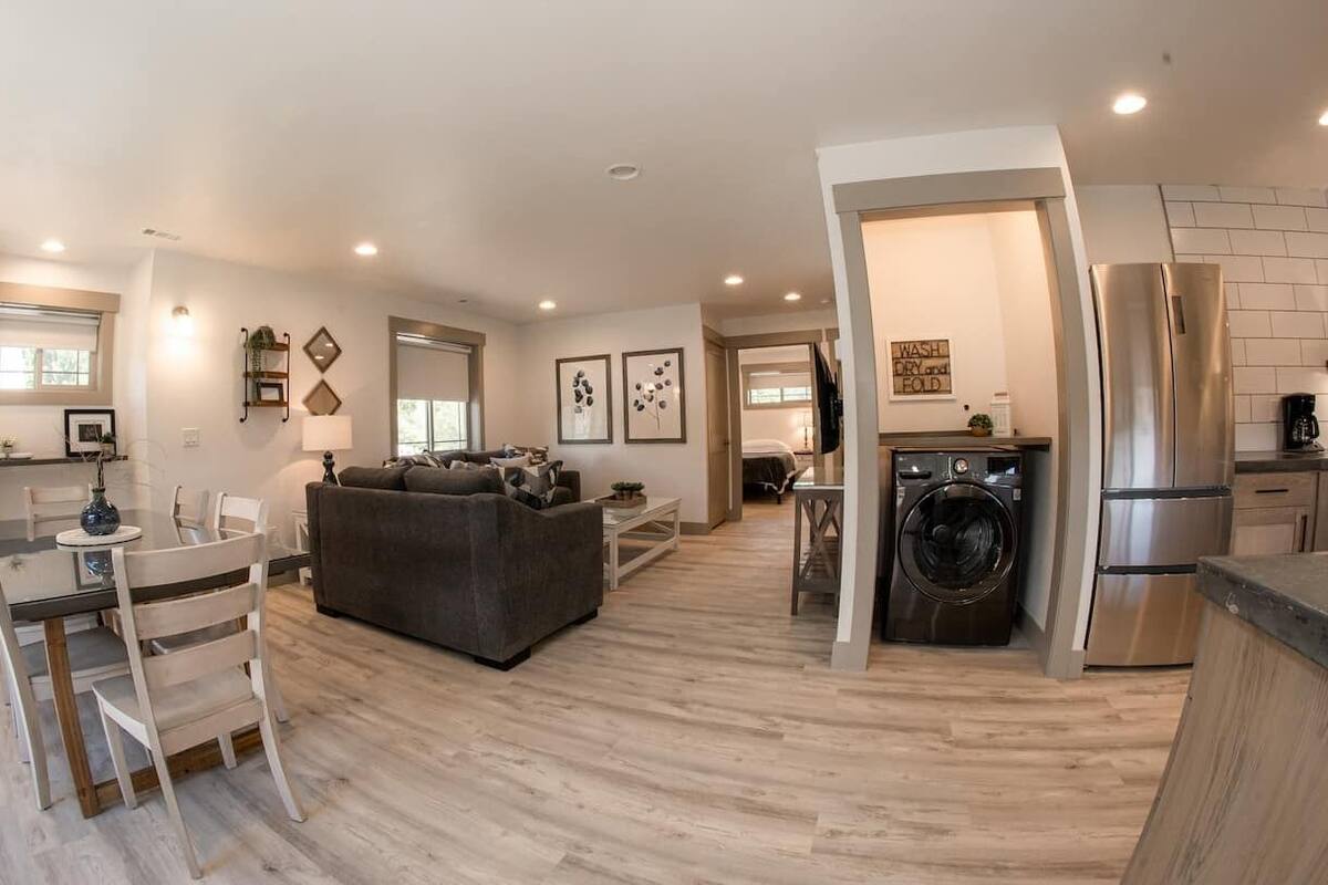 A modern kitchen with stainless steel appliances, a dining area with a white table and chairs, and a living room with a gray sofa.