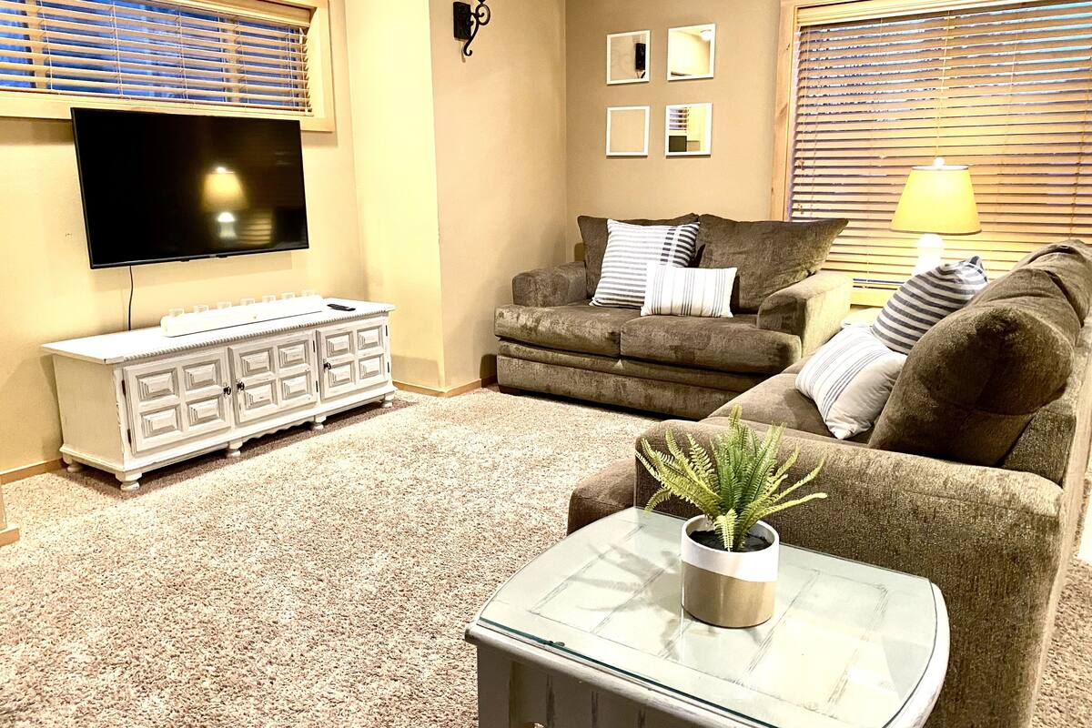A living room with a flat-screen TV, a white entertainment unit, two couches, a glass coffee table with a plant, and a lamp.