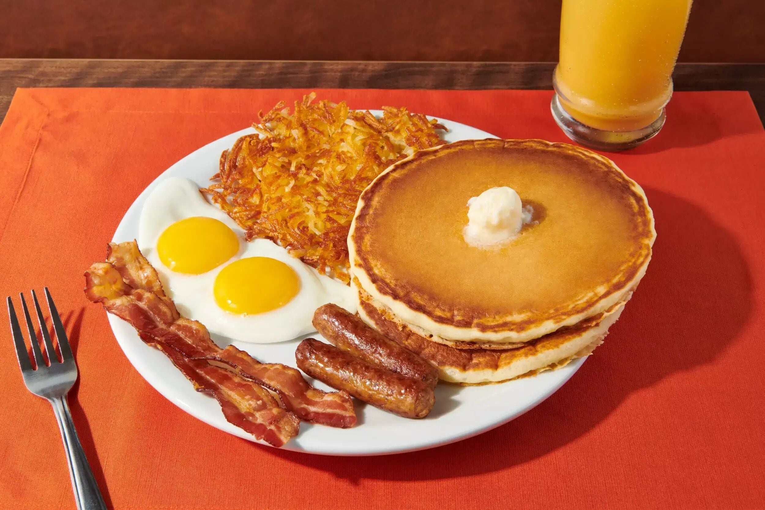 A breakfast plate with eggs, bacon, hash browns, pancakes, and a glass of orange juice.
