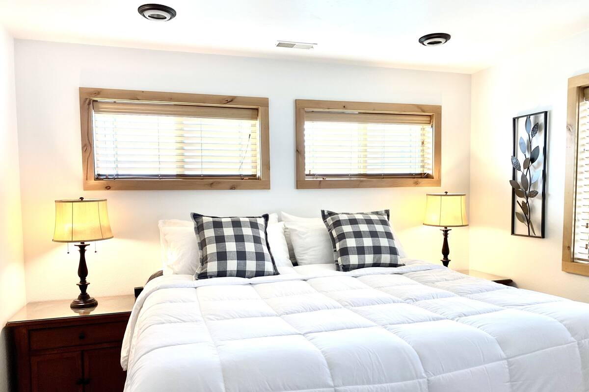 A bedroom with a large bed, two bedside tables with lamps, and two windows with blinds.