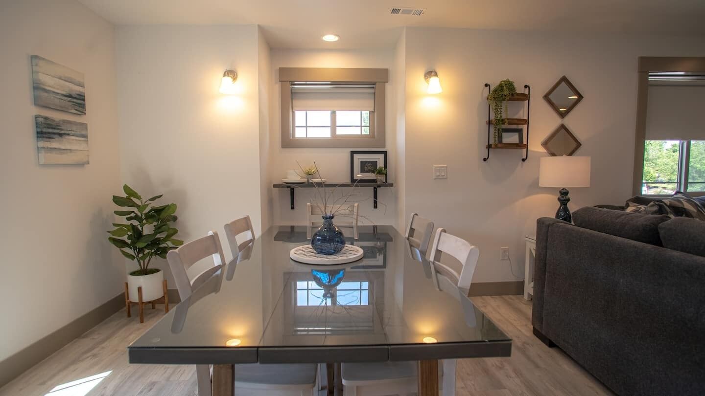 A modern dining area with a glass table, white chairs, and a vase centerpiece. There is a grey sofa and a small plant in the corner.