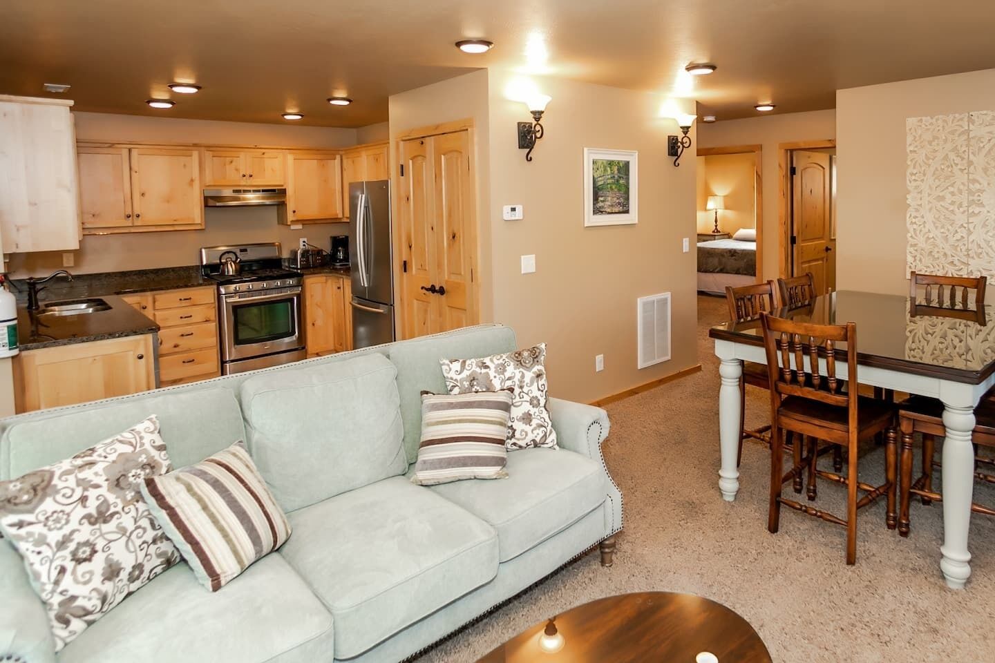 A modern kitchen with wooden cabinets and stainless steel appliances, a light-colored sofa with patterned cushions, and a dining area with a wooden table and chairs.