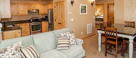 A modern kitchen with wooden cabinets and stainless steel appliances, a light-colored sofa with patterned cushions, and a dining area with a wooden table and chairs.